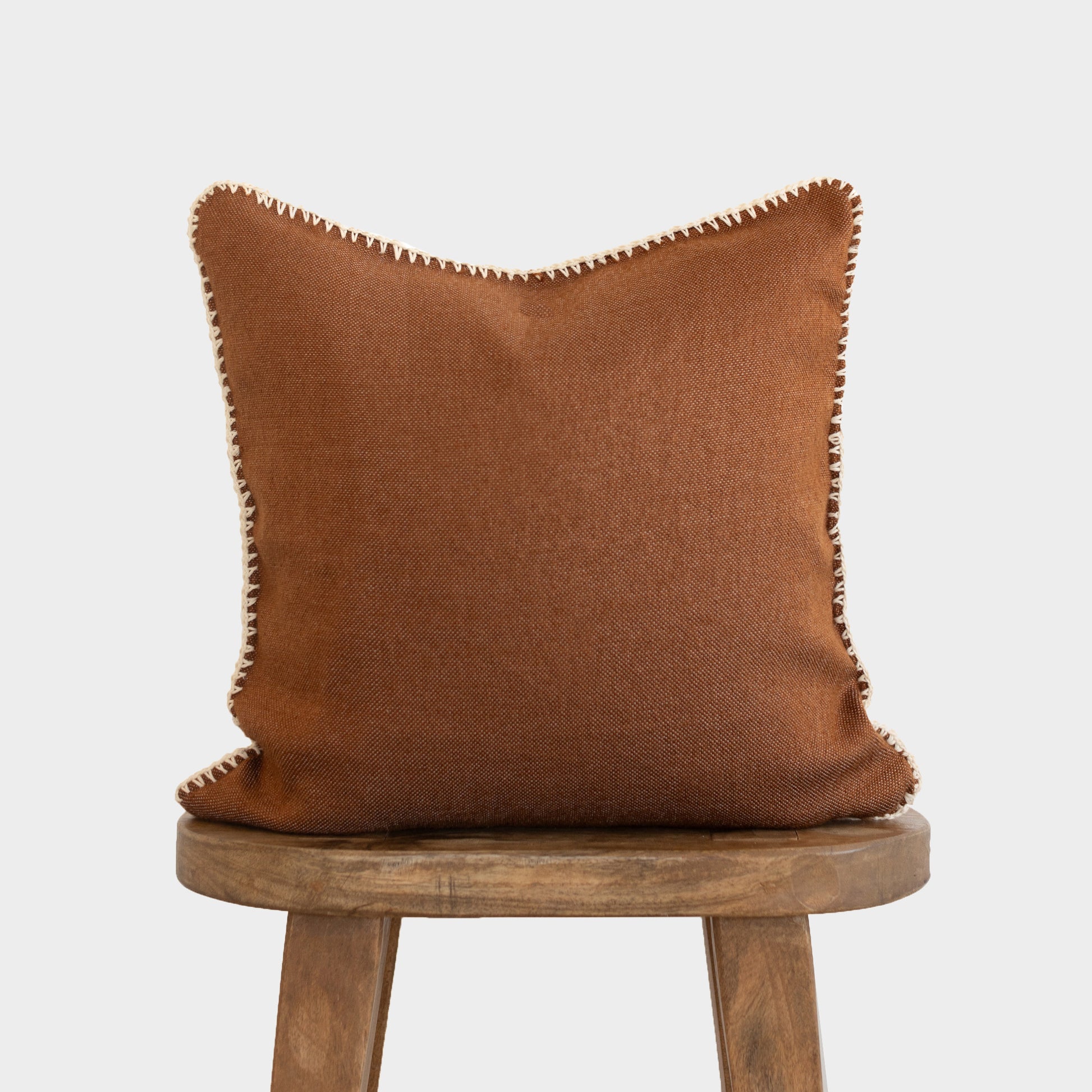 Rust-brown cushion with whipstitched edges displayed on a wooden stool.