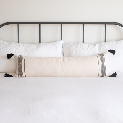 Cream lumbar pillow with gray striped trim and black tassels displayed on a bed with white pillows and black metal headboard.