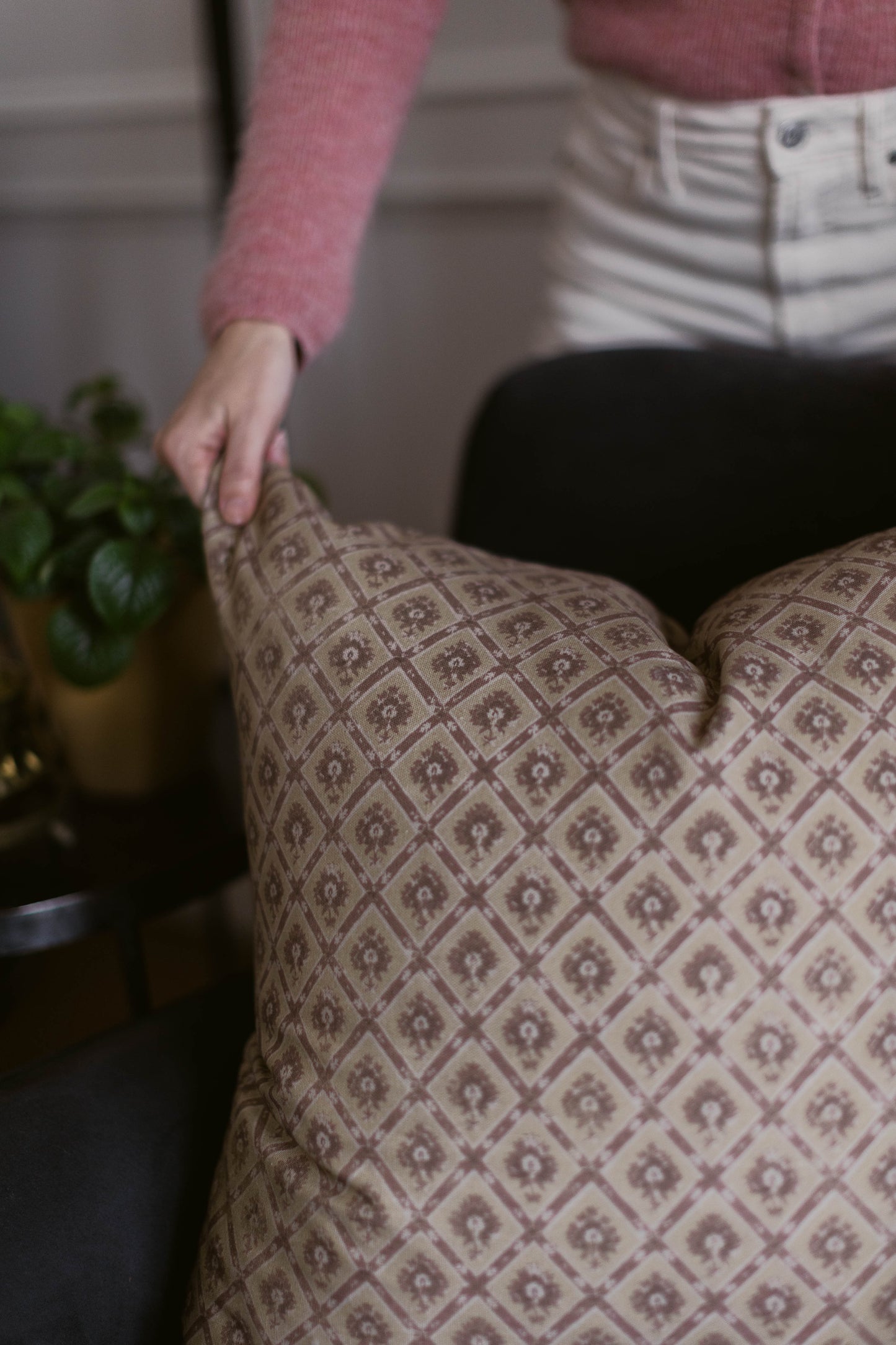 Beige decorative pillow with geometric floral pattern, displayed on furniture with hand touching surface.