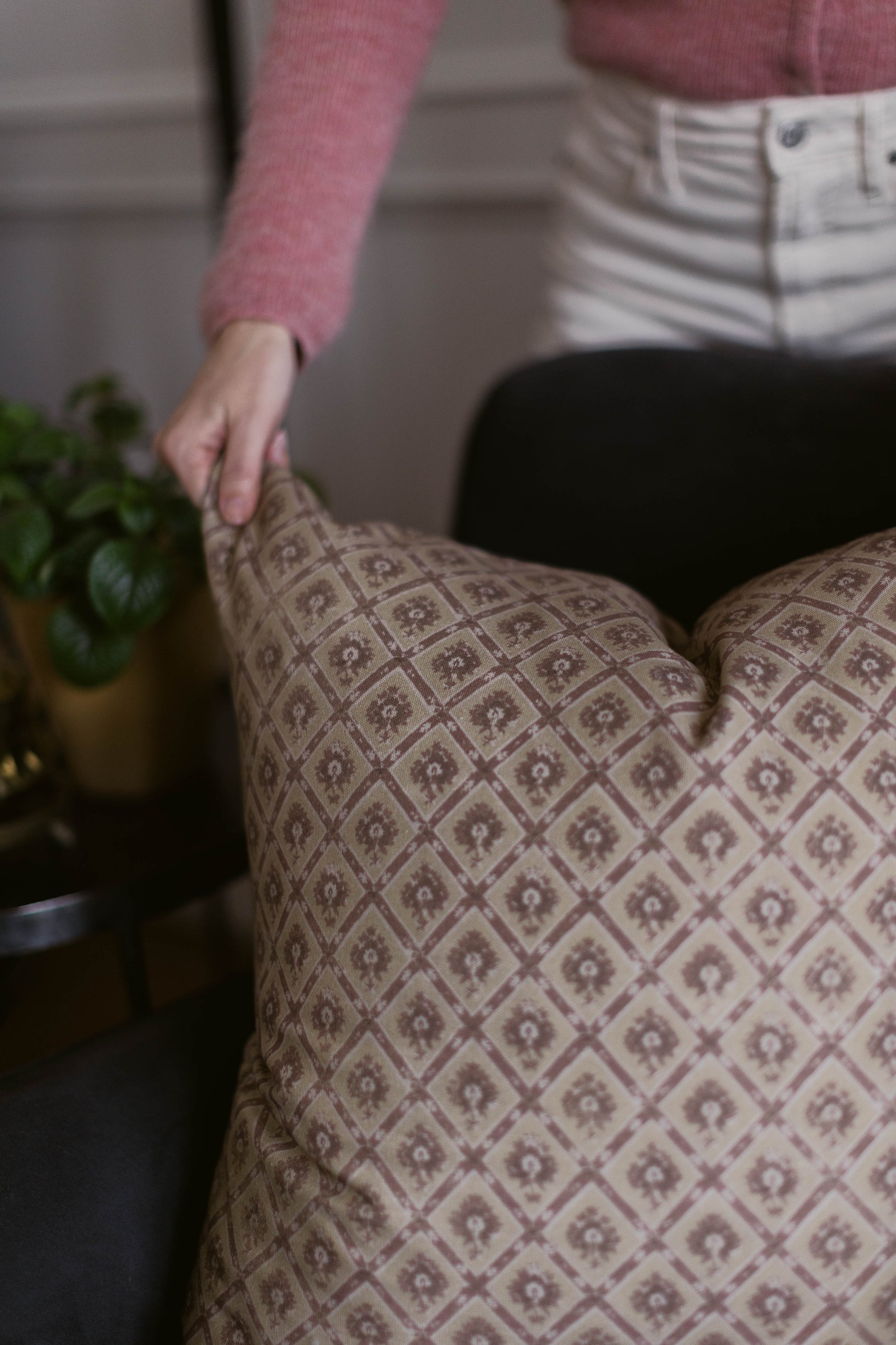 Beige decorative pillow with geometric floral pattern, displayed on furniture with hand touching surface.