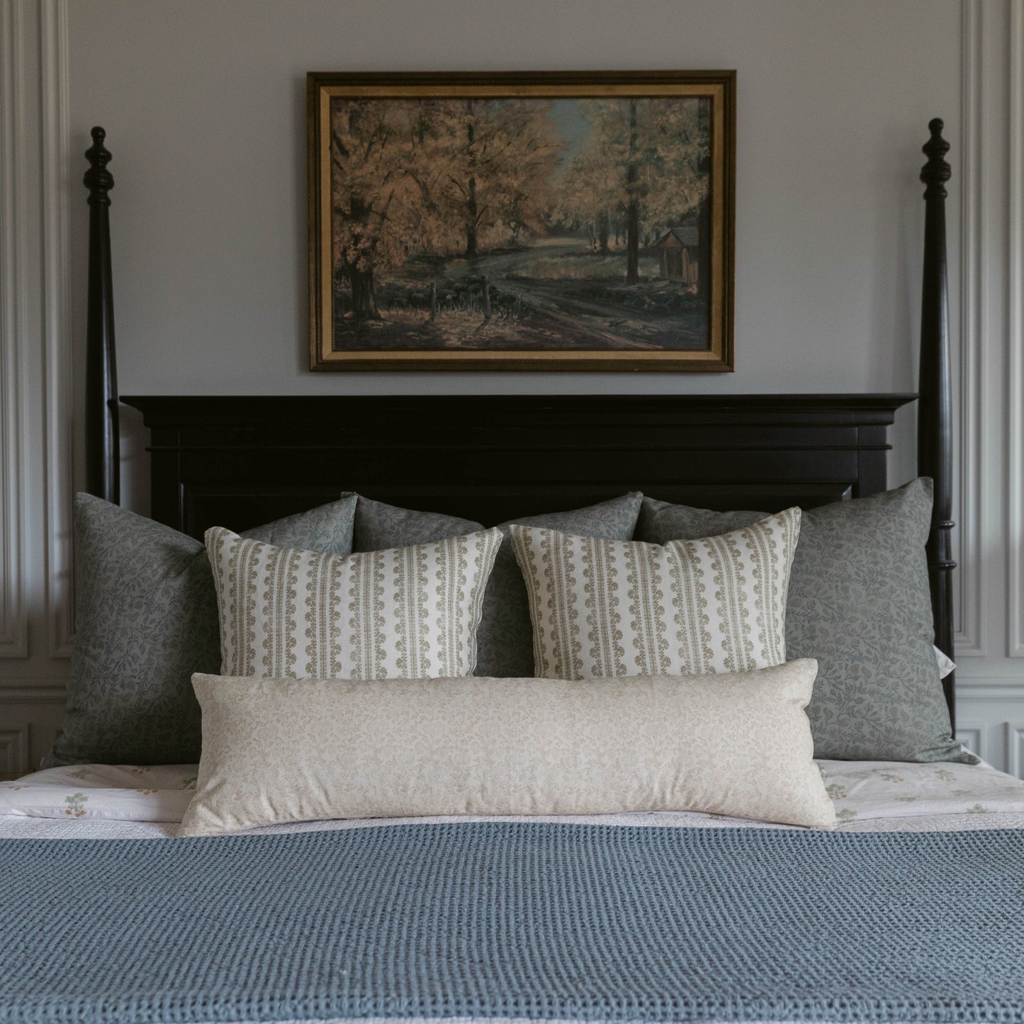 Framed landscape artwork with gold border hanging above a black four-poster bed styled with patterned and solid linen pillows