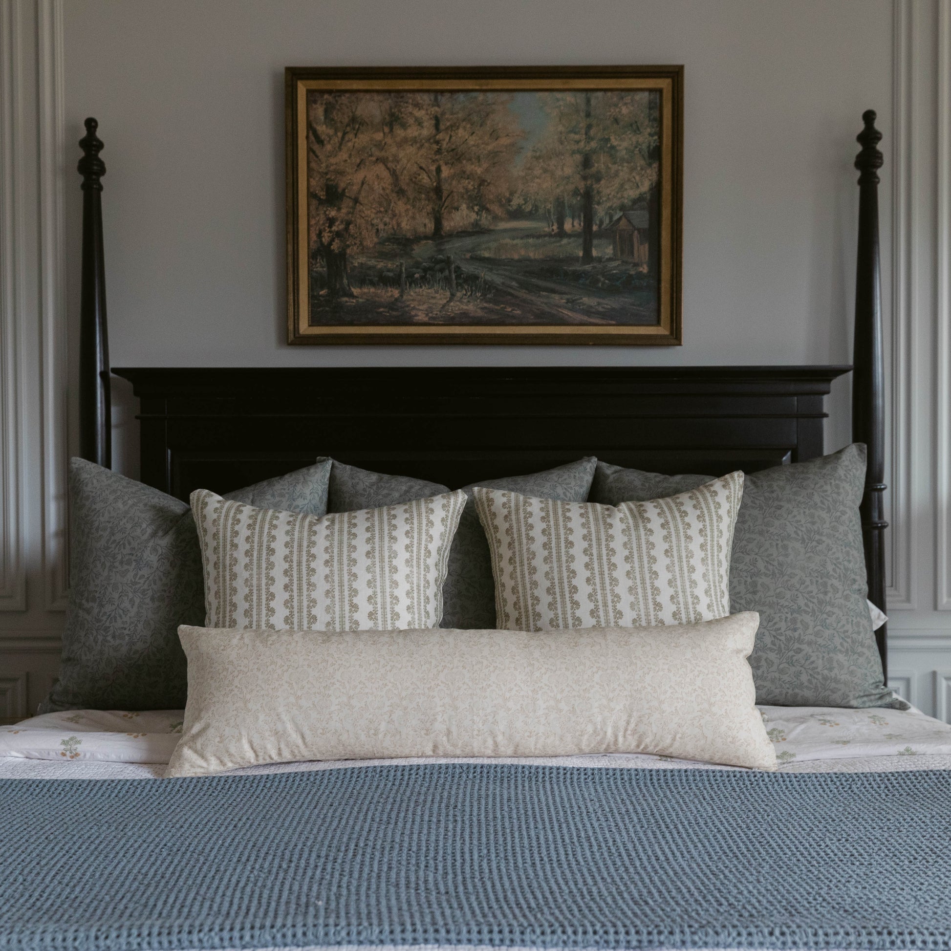 Framed landscape artwork with gold border hanging above a black four-poster bed styled with patterned and solid linen pillows
