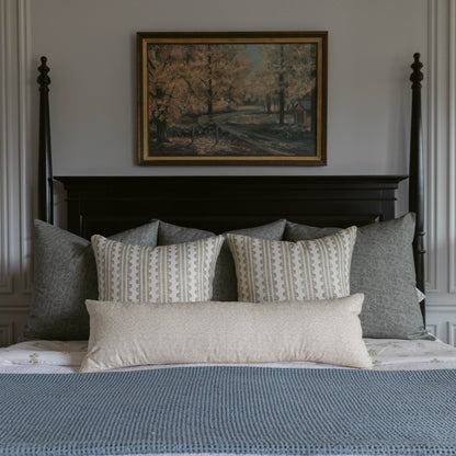 Framed landscape artwork with gold border hanging above a black four-poster bed styled with patterned and solid linen pillows