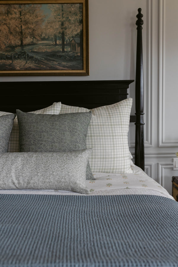 Grey and cream linen pillow with floral pattern displayed on bed with plaid and striped bedding.