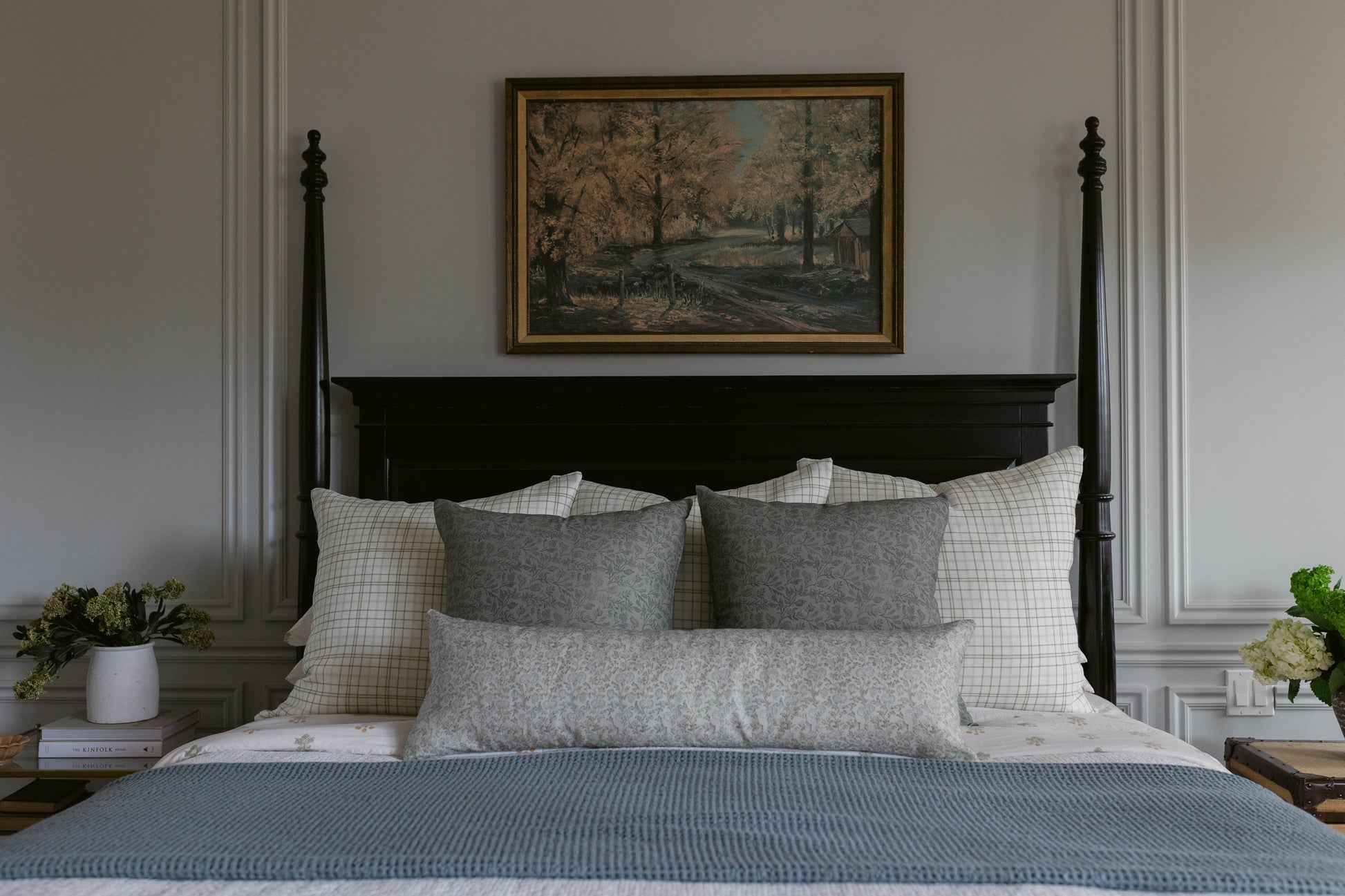 Framed landscape painting with gold border hanging above black four-poster bed with gray and white bedding.