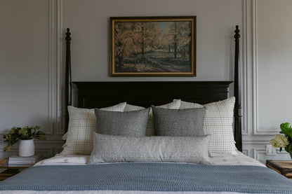 Framed landscape painting with gold border hanging above black four-poster bed with gray and white bedding.