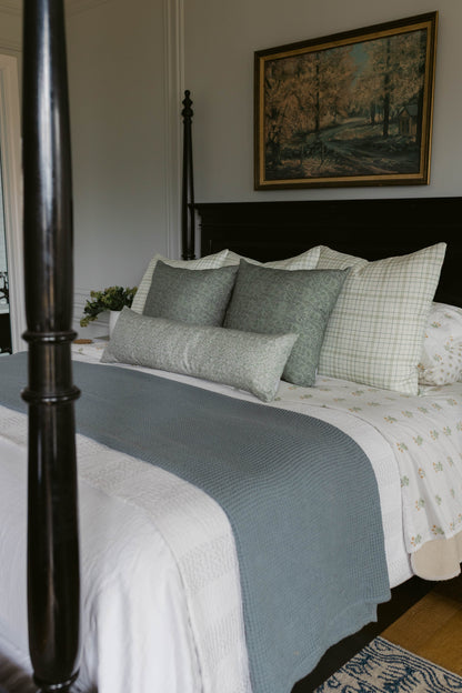 Linen pillows in sage green and cream grid patterns arranged on a made bed with blue throw blanket.