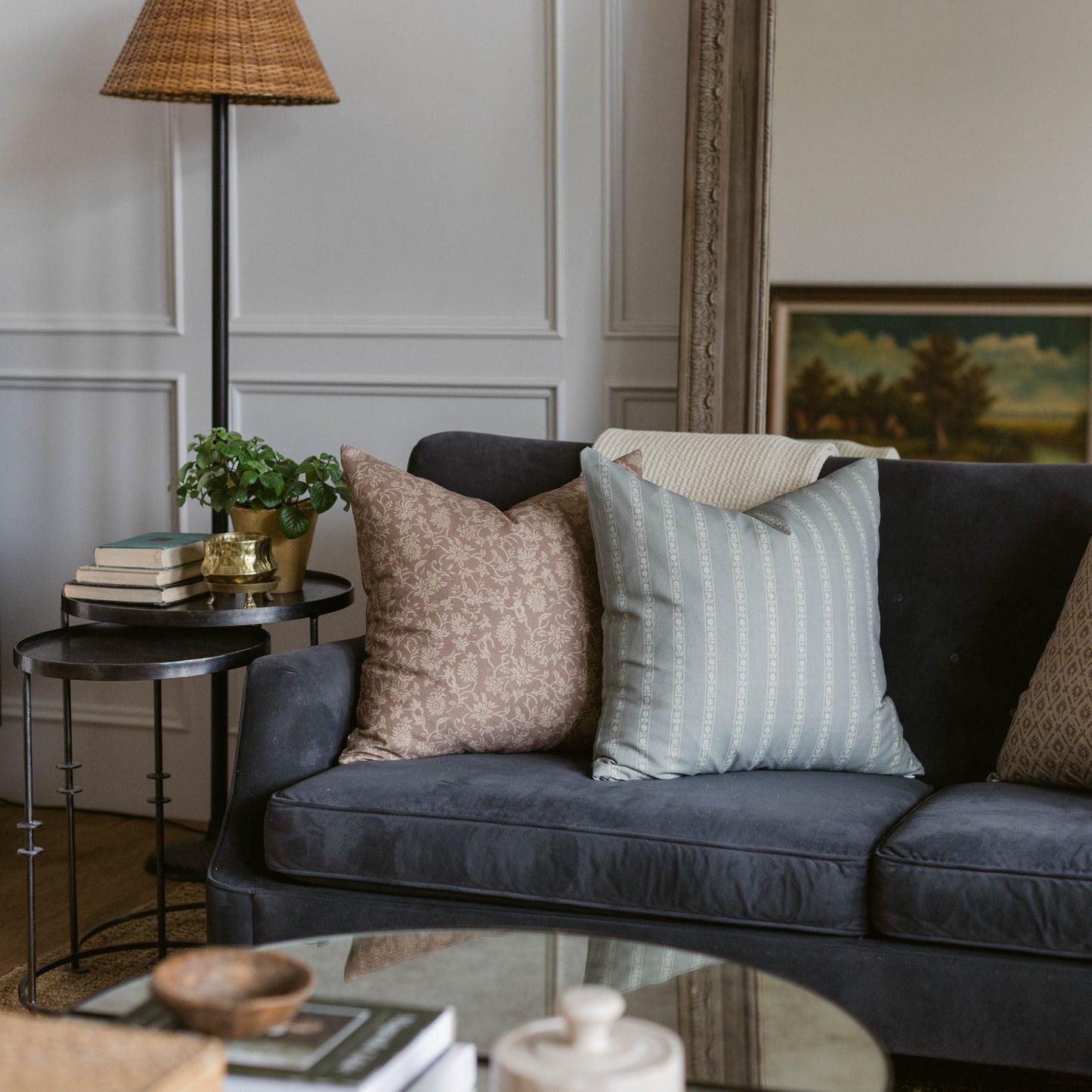 Woven rattan cone lamp with black stem on dark sofa with patterned throw pillows.