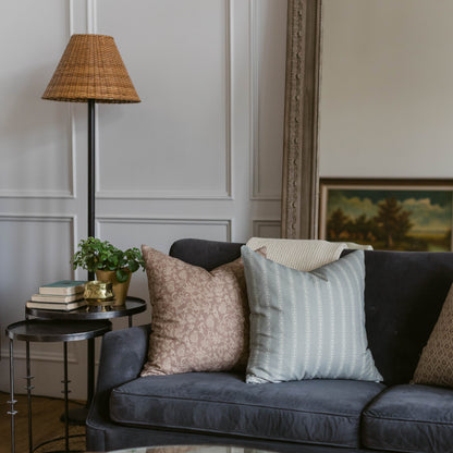 Woven wicker cone-shaped lampshade in tan with black pole stem displayed above dark upholstered sofa with decorative pillows.
