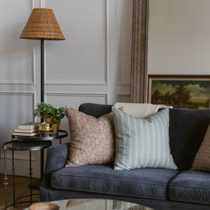 Woven wicker trapezoid lamp shade in tan with dark metal pole, displayed on side table next to navy sofa with decorative pill