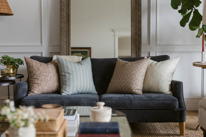 Noa throw pillow in taupe displayed on dark navy sofa with striped and textured accent pillows in living room setting.