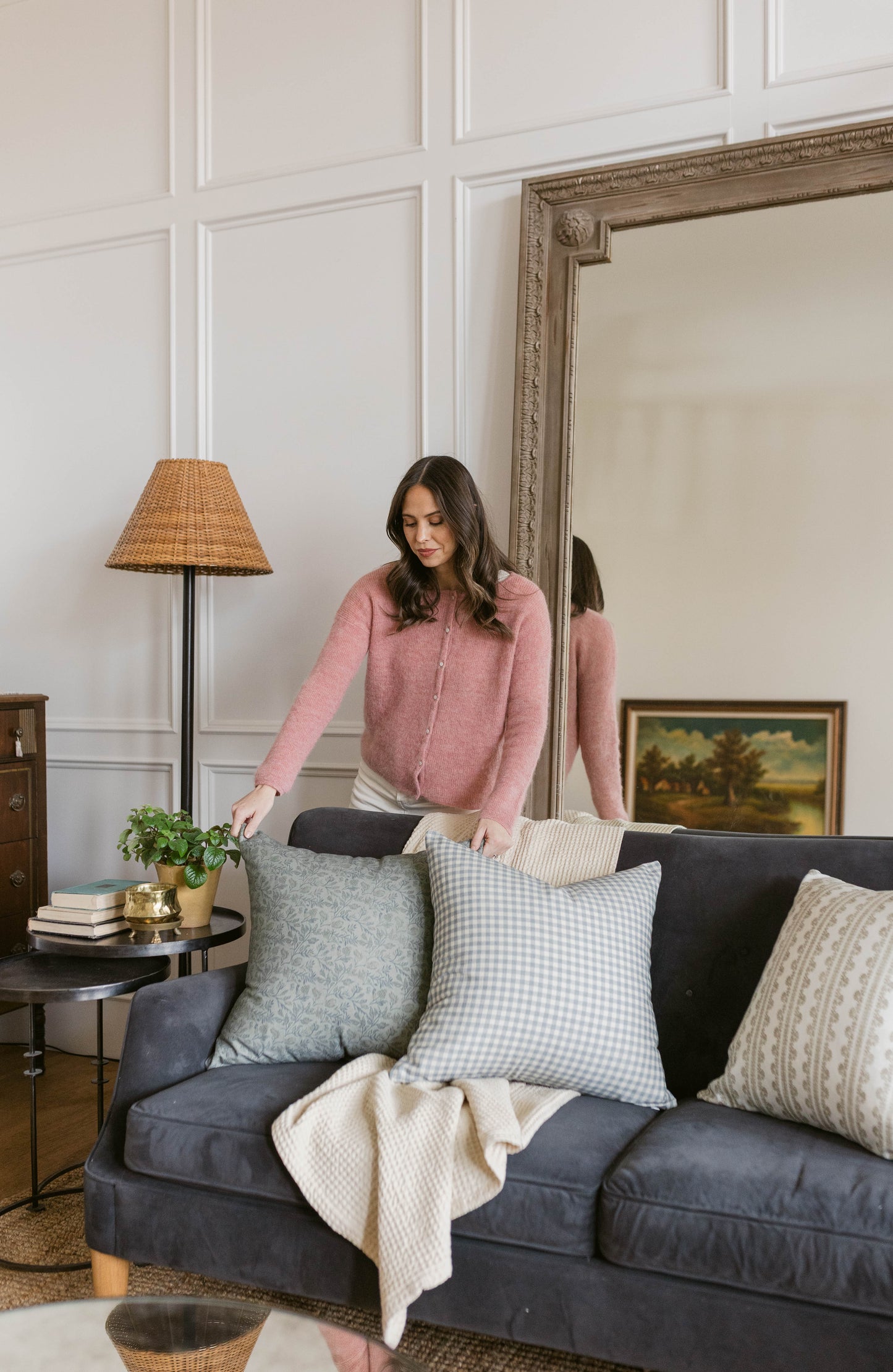 Woman wearing pink button-up cardigan standing beside dark sofa with patterned throw pillows.