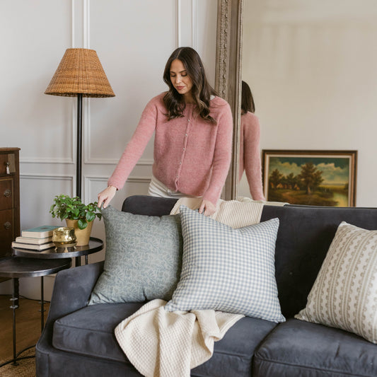 Dusty blue and white gingham check pillow on dark sofa with woman arranging decor.