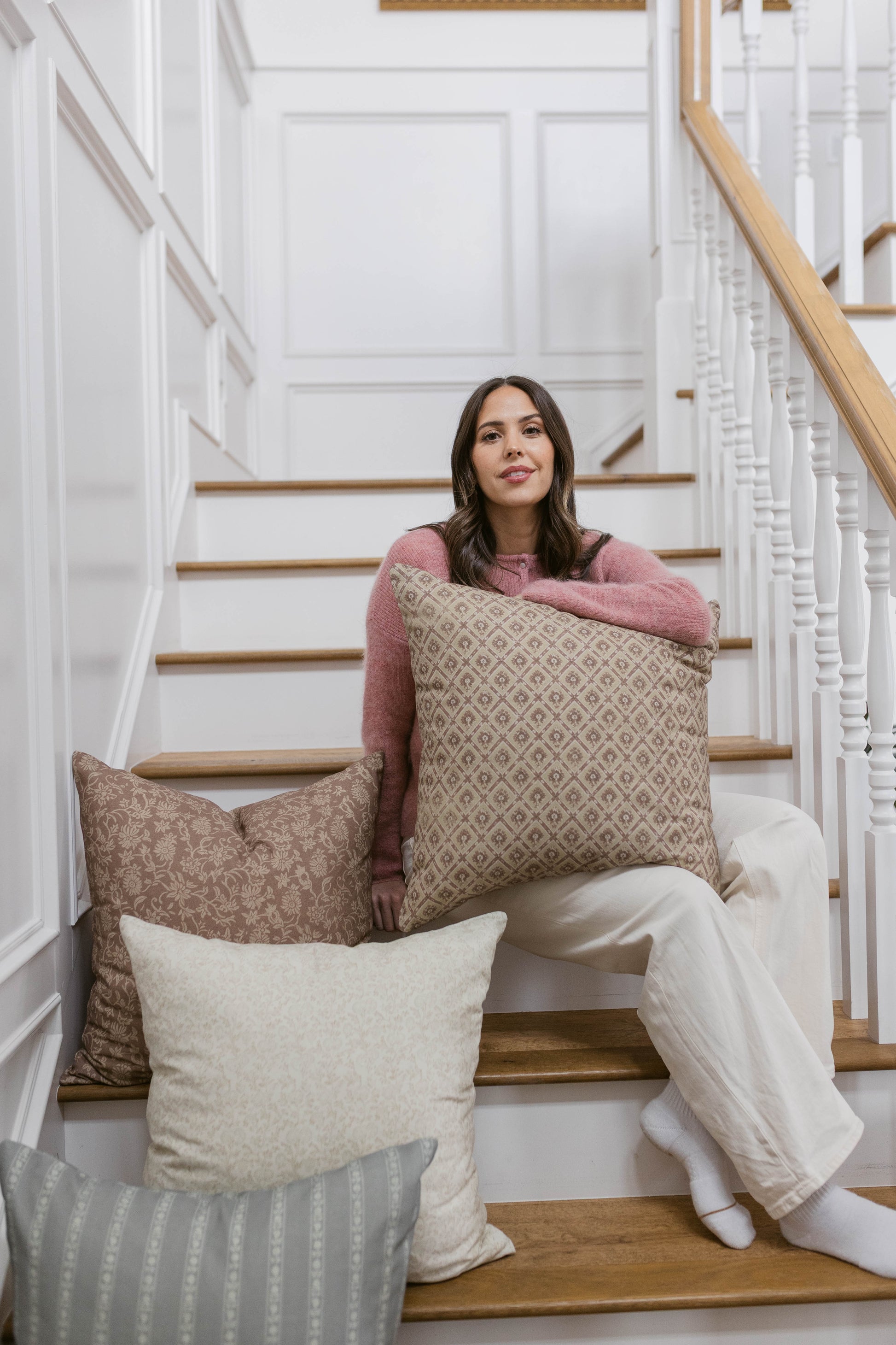 Woman holding beige geometric patterned pillow on staircase with additional decorative pillows.