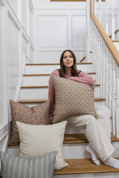 Woman holding beige geometric patterned pillow on staircase with additional decorative pillows.