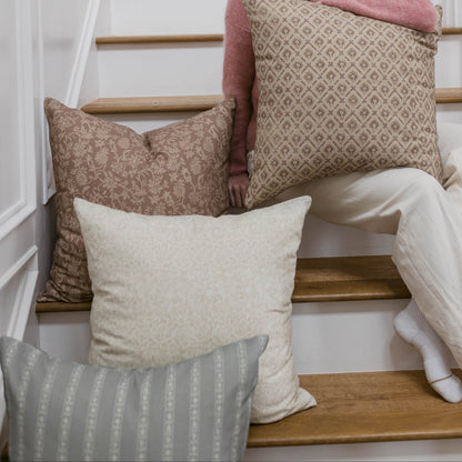 Multiple decorative pillows in taupe, cream, and pink tones with geometric and floral patterns arranged on wooden stairs.