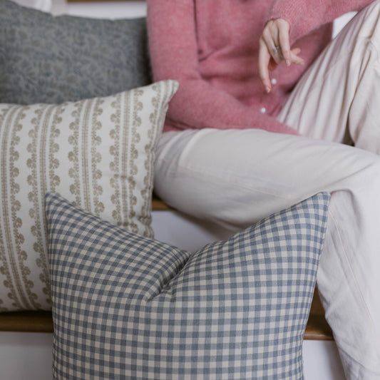 Four decorative pillows in cream, blue gingham, gray, and pink displayed on a couch.