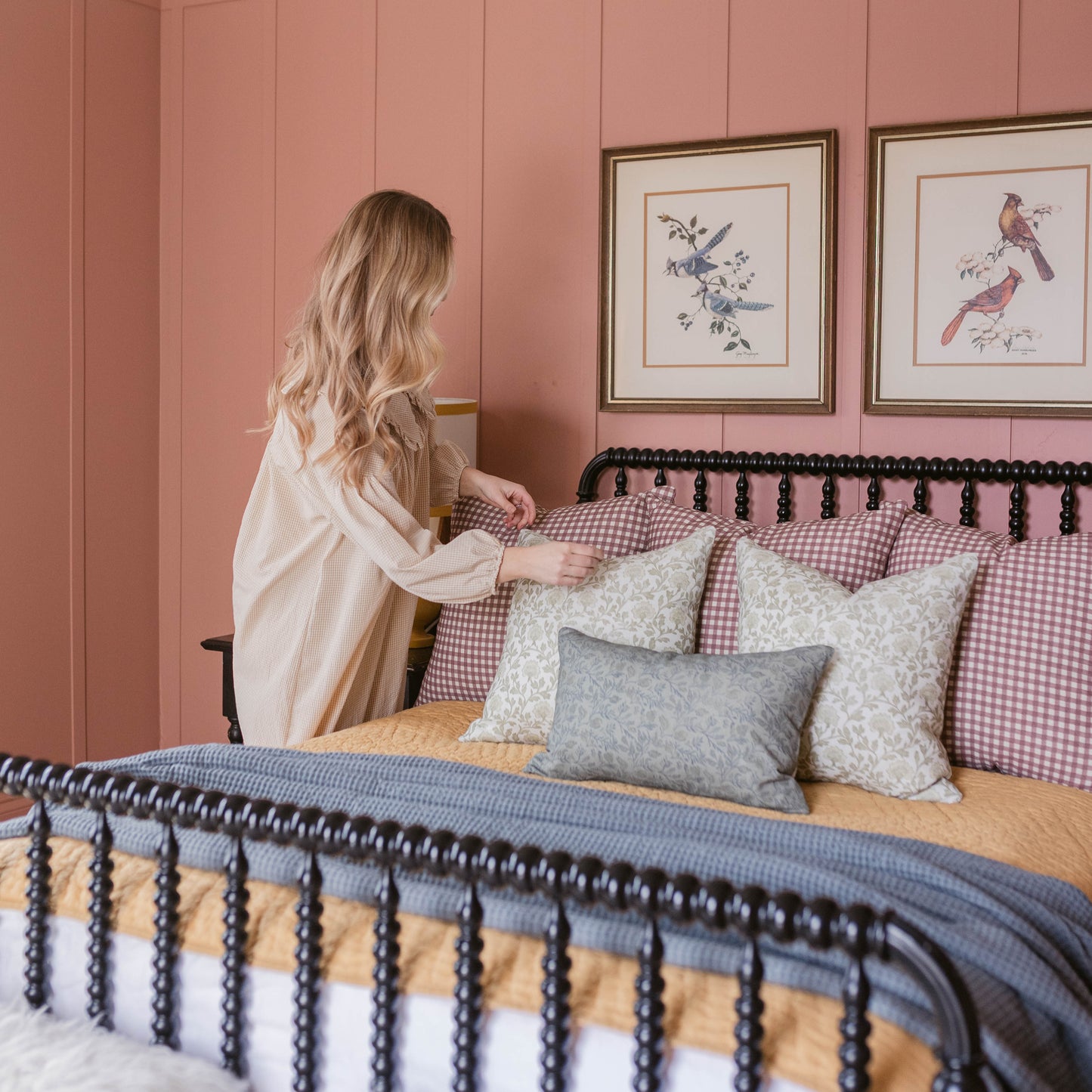 Woman in white shirt sits on bed with blue bedding and patterned pillows against pink wall with framed bird artwork.