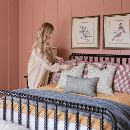 Woman in white shirt sits on bed with blue bedding and patterned pillows against pink wall with framed bird artwork.