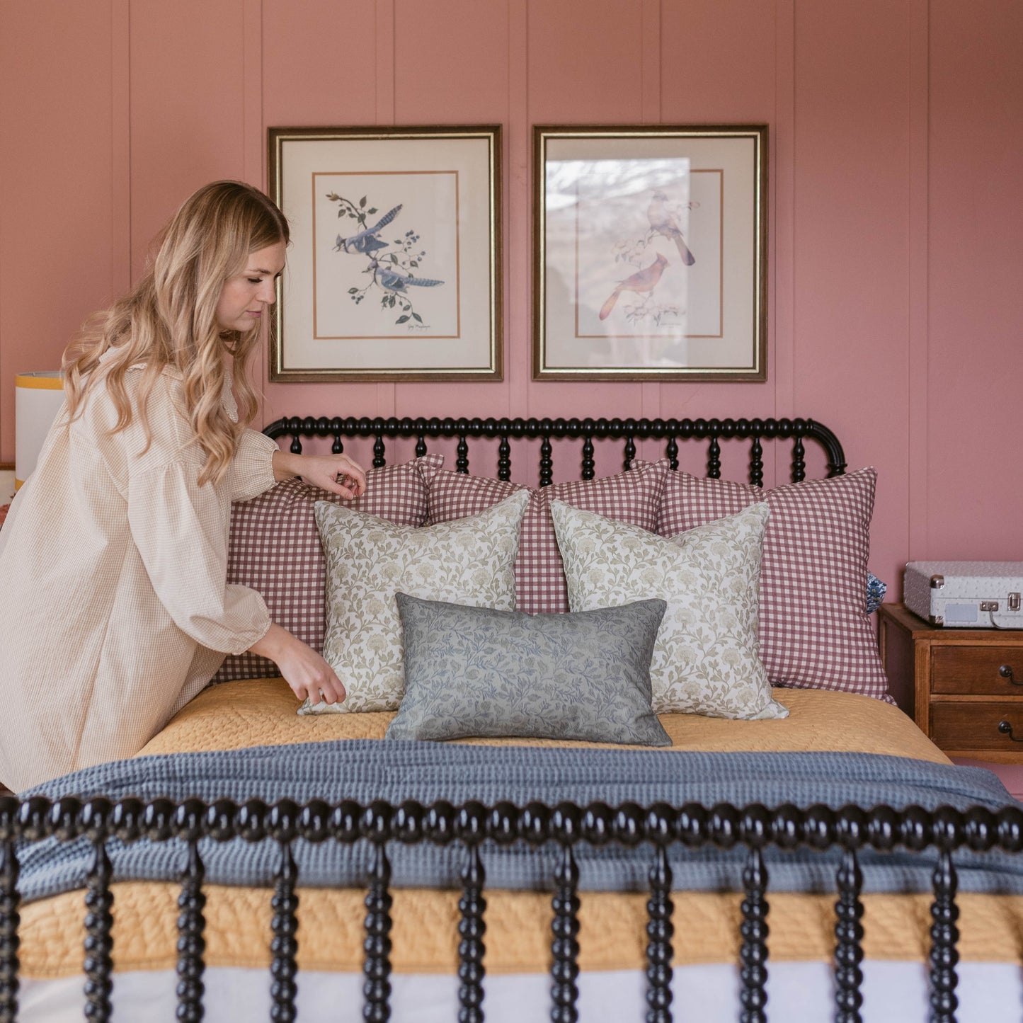 Woman in cream cardigan adjusting patterned pillows on bed with black tassel trim headboard against pink wall.