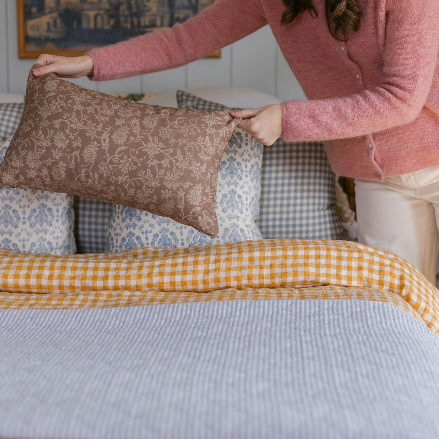 Person holding brown damask patterned rectangular pillow on bed with yellow gingham and blue patterned bedding.