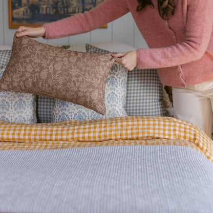 Person holding brown damask patterned rectangular pillow on bed with yellow gingham and blue patterned bedding.