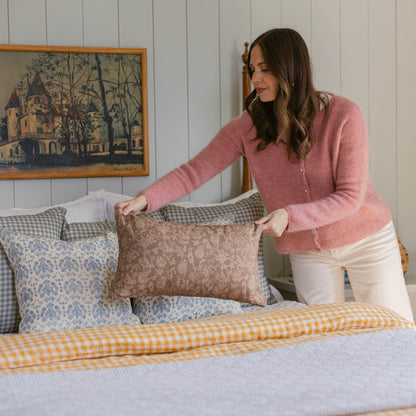 Woman arranging patterned pillows on a bed with yellow gingham bedding and framed castle artwork on wall.