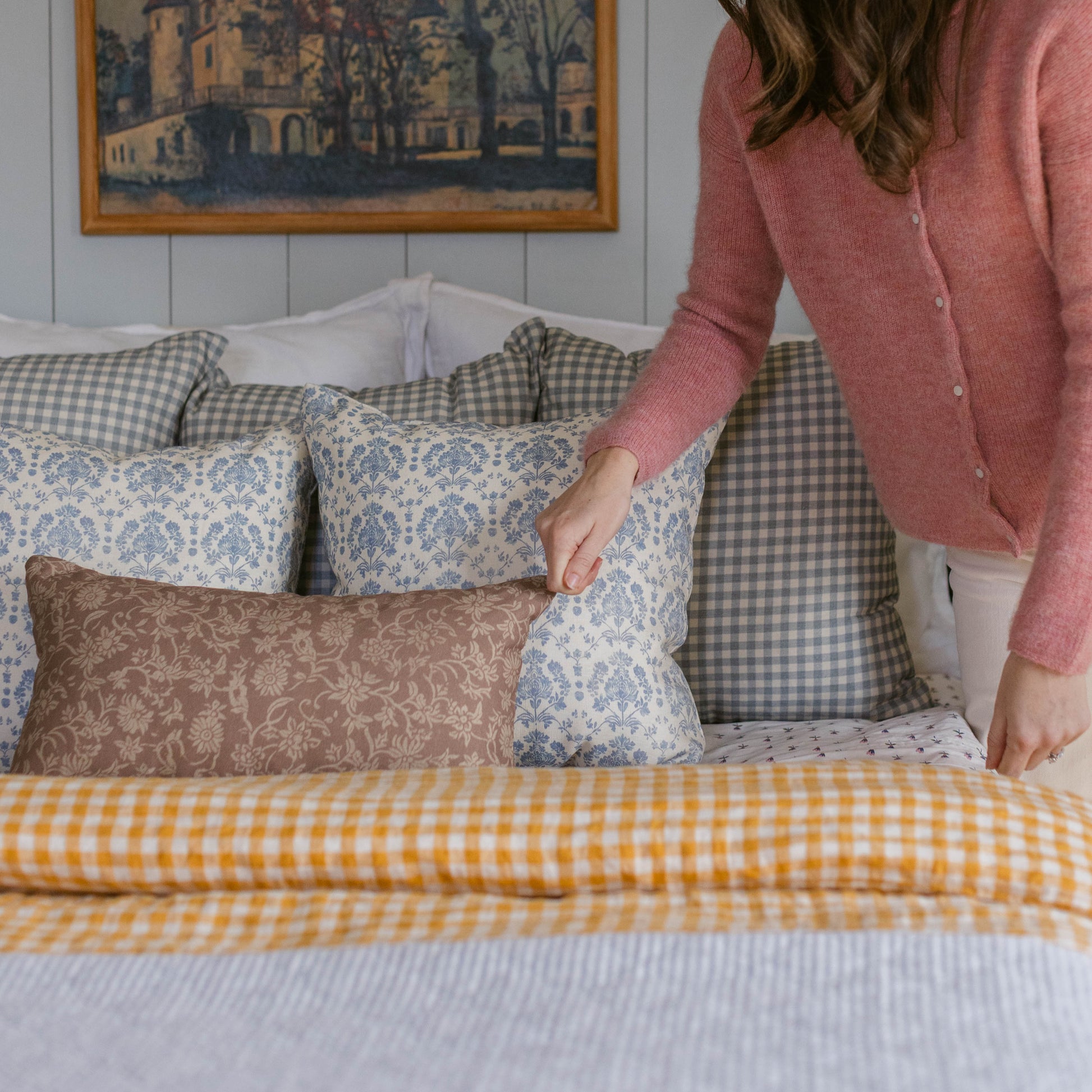 Person arranging decorative pillows on bed with yellow gingham duvet and patterned throw pillows.