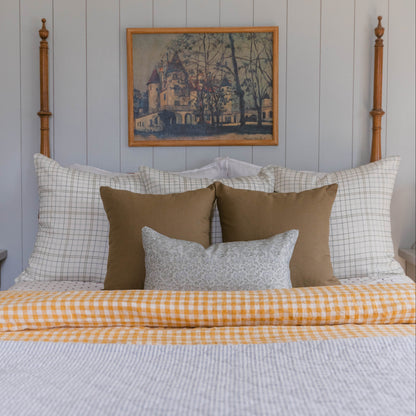 Bed with wooden finial posts, white grid pillows, brown accent pillows, gray textured pillow, and yellow gingham coverlet.