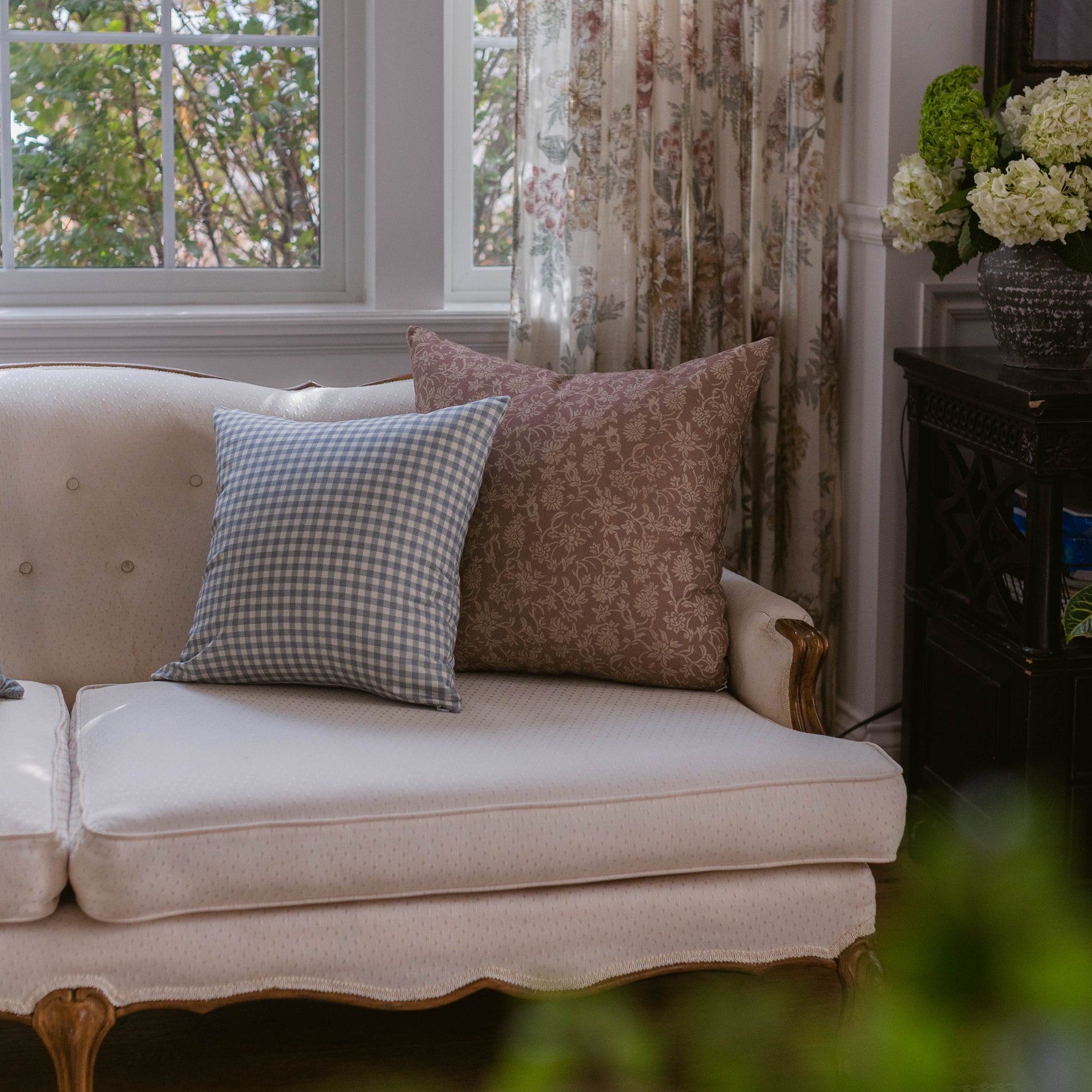Two decorative pillows on a cream upholstered sofa: one with blue gingham pattern and one with brown floral damask design.