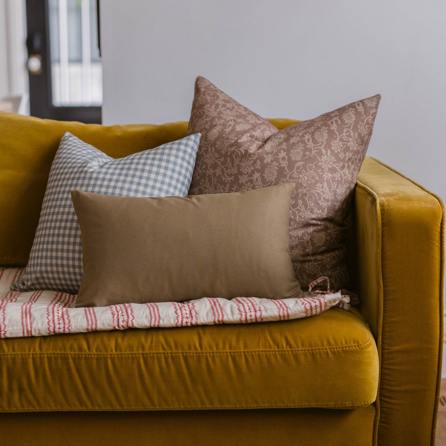 Three decorative pillows on a mustard-yellow sofa: a blue and white gingham pillow, a solid tan pillow, and a brown damask pi
