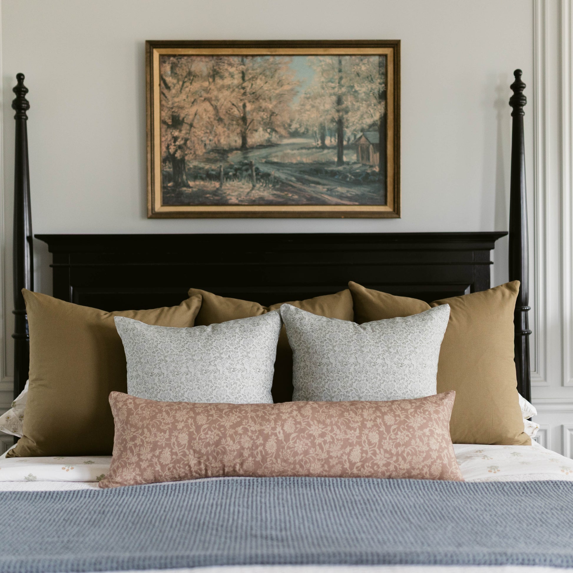 Framed landscape artwork with vintage forest and water scene in gold and black frame mounted above bed with pillows.