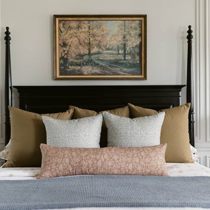 Framed landscape artwork with vintage forest and water scene in gold and black frame mounted above bed with pillows.