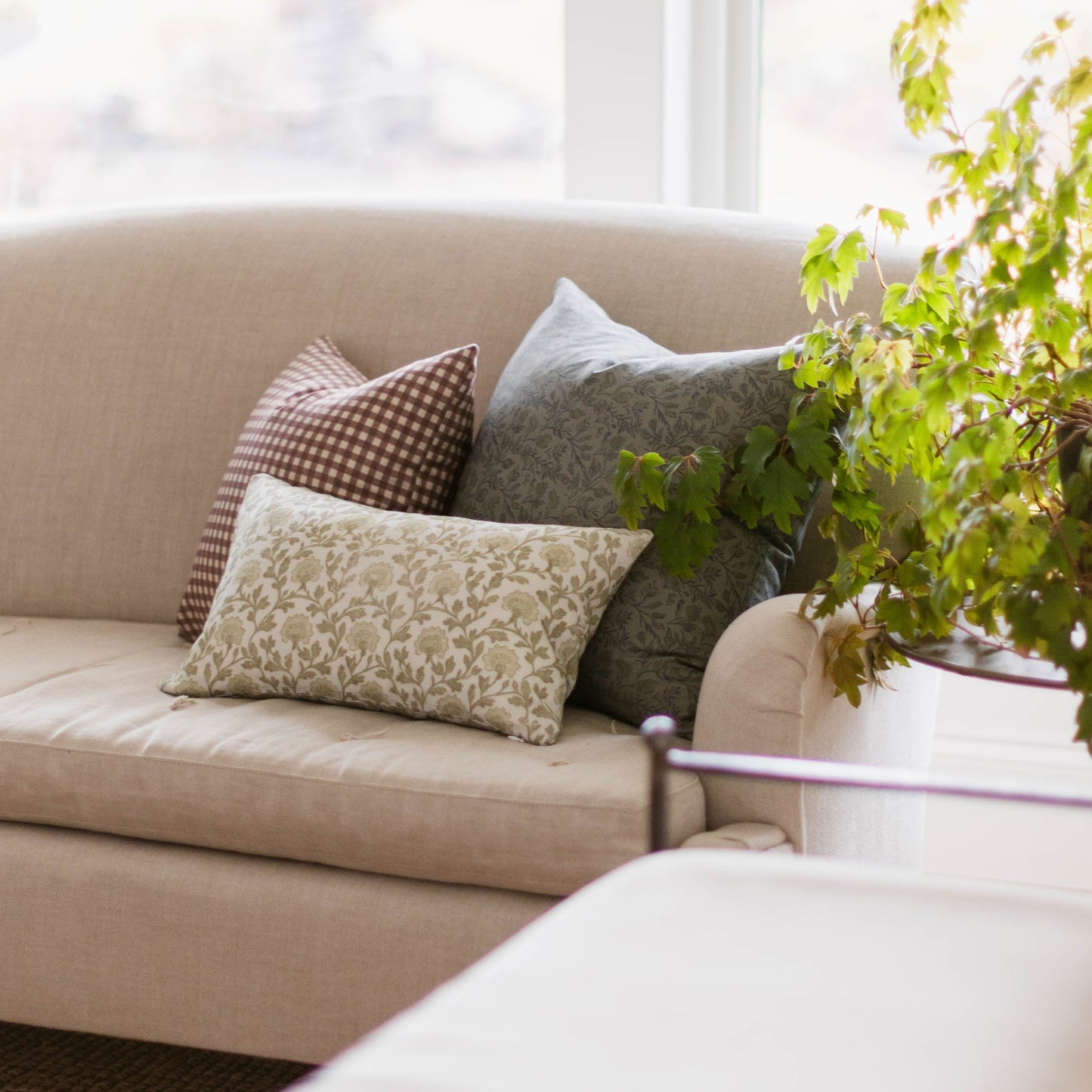 Beige linen pillow with subtle leaf pattern displayed on cream sofa with coordinating decorative pillows near a window.