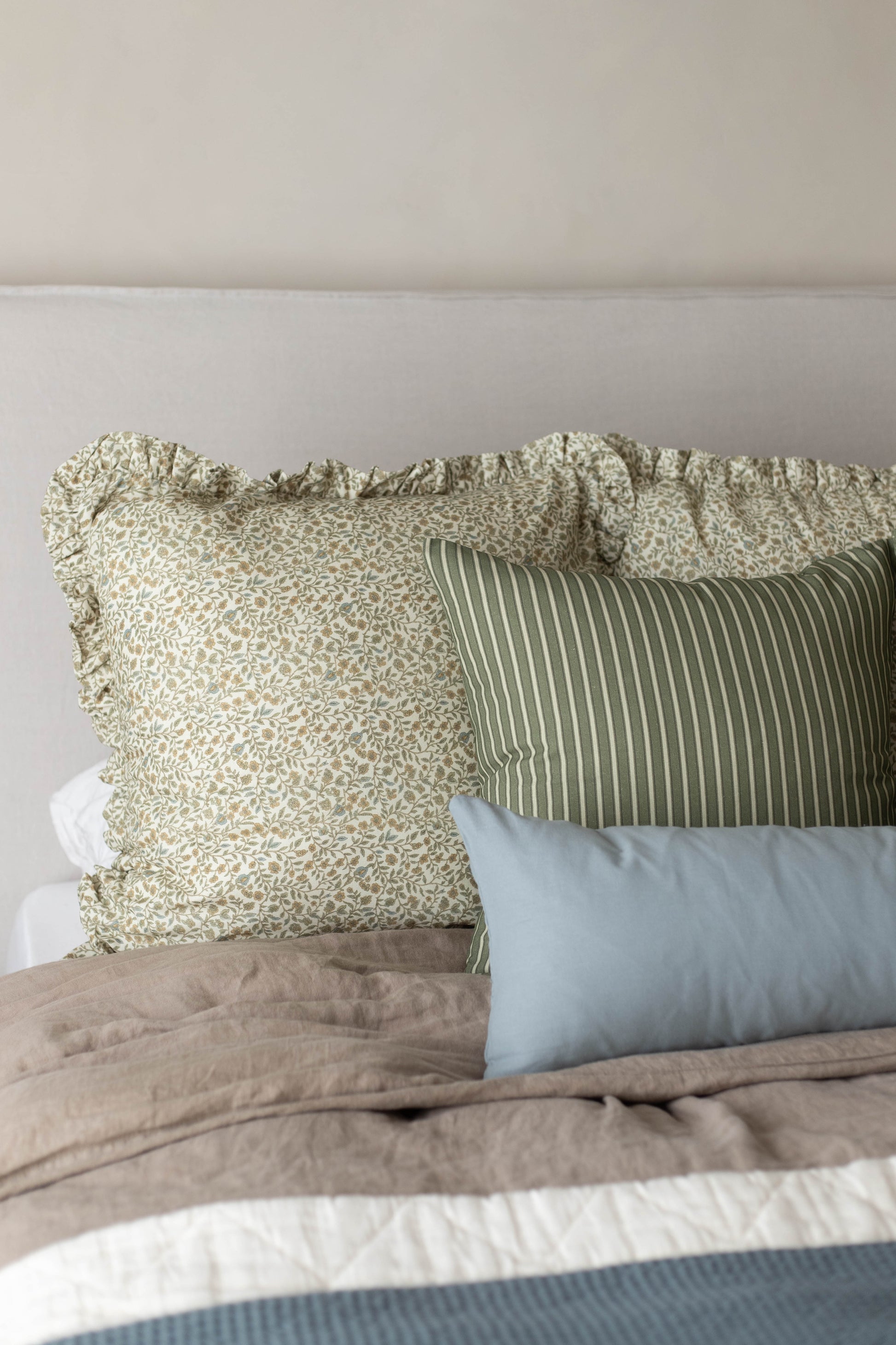 Bed with dusty blue pillow and patterned throw pillows in cream and sage green with ruffled edges.