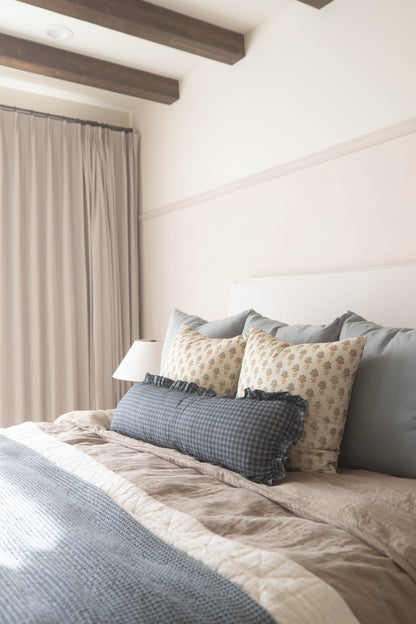 Dusty blue gingham lumbar pillow with ruffled edges displayed on a bed with patterned pillows.