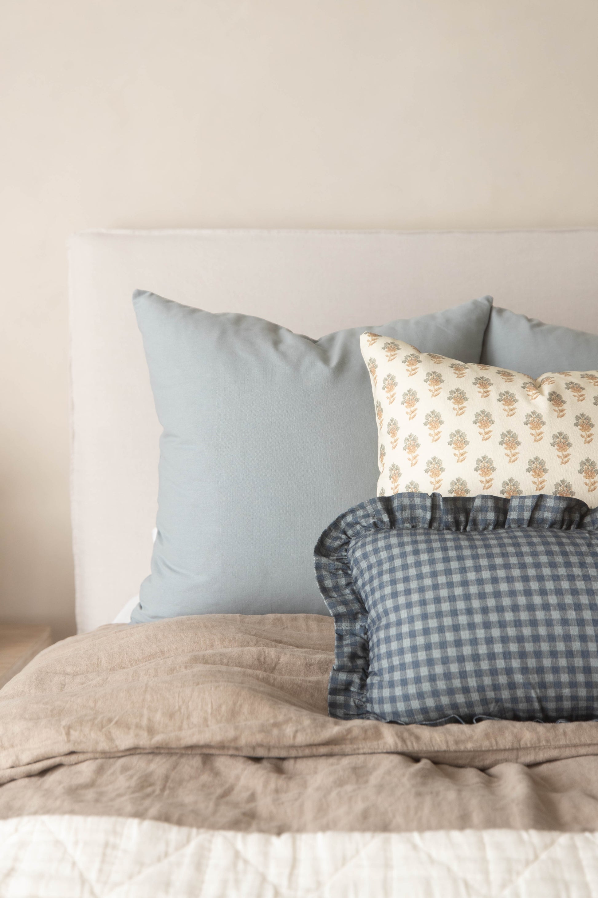 Dusty blue solid pillow displayed with patterned and gingham pillows on a bed with beige bedding.