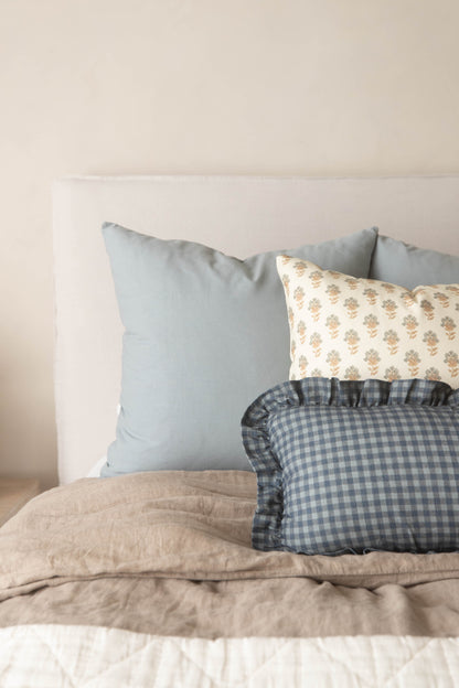 Dusty blue solid pillow displayed with patterned and gingham pillows on a bed with beige bedding.