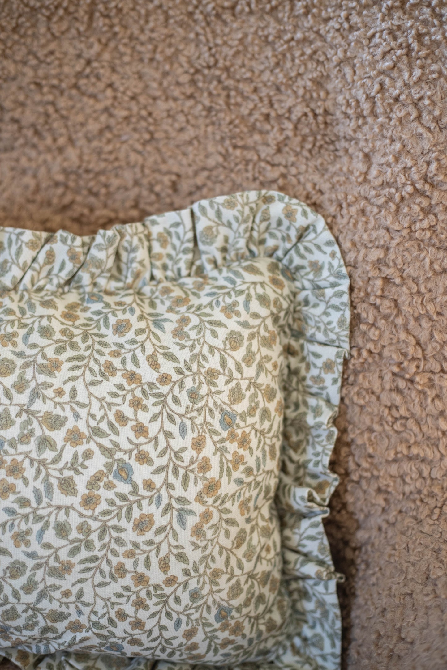 Cream pillow with ruffled edge and gold-green leaf print pattern displayed against textured beige fabric.