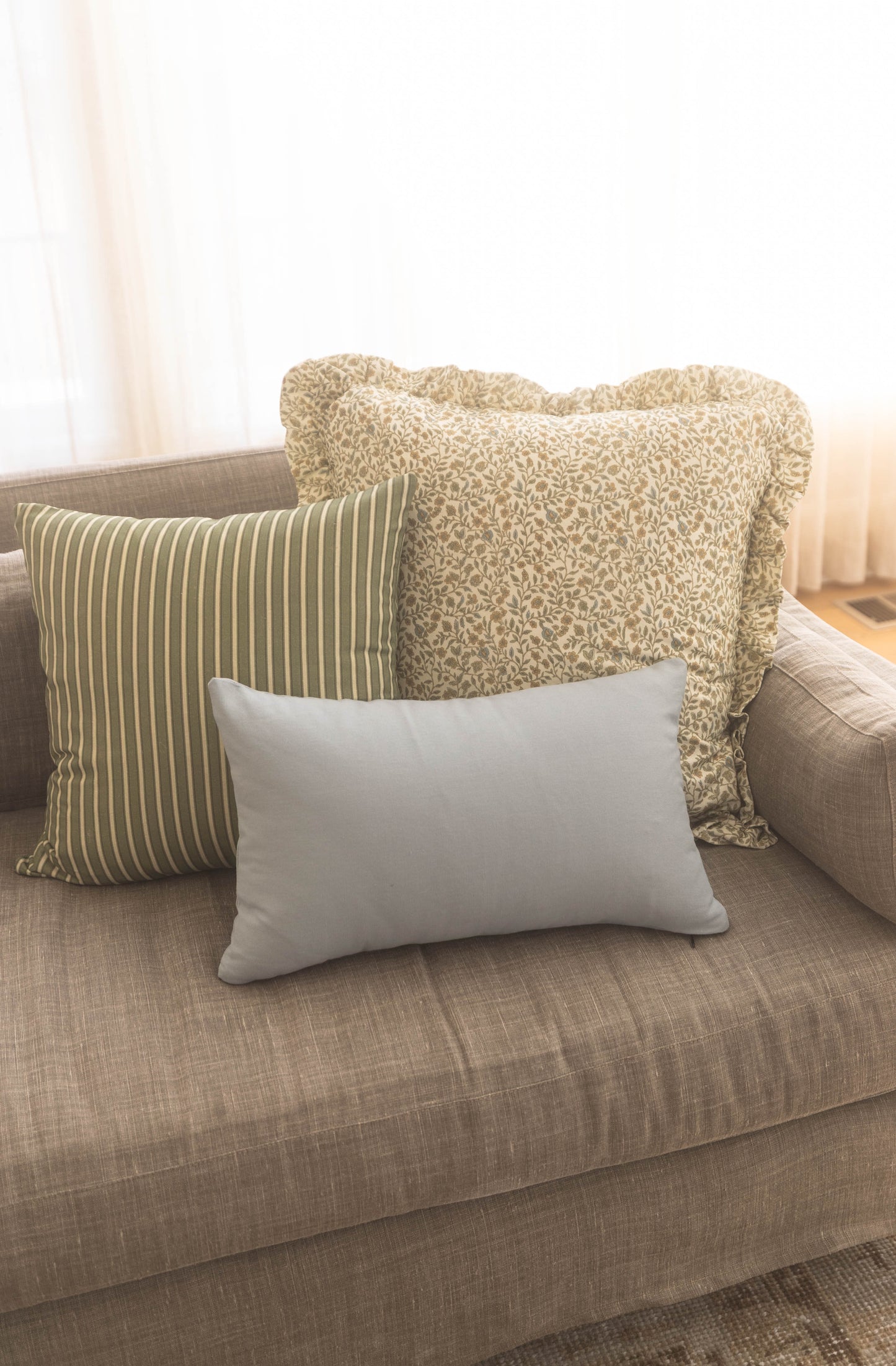Three throw pillows on a beige sofa: striped green and cream, textured cream with ruffled edges, and solid gray rectangular p