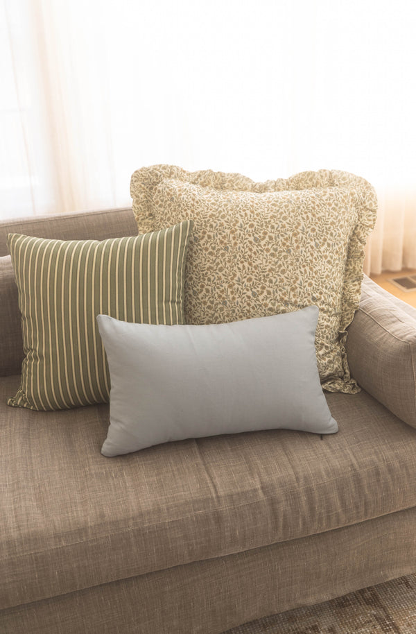 Three throw pillows on a beige sofa: striped green and cream, textured cream with ruffled edges, and solid gray rectangular p
