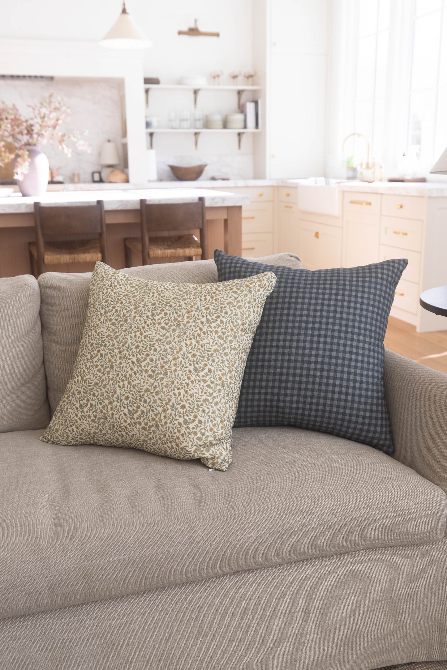 Two decorative pillows on a beige sofa: one with cream leaf pattern, one with charcoal gingham check.
