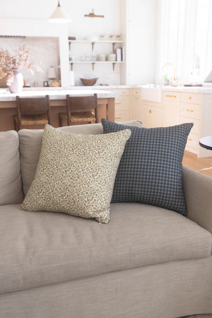 Two decorative pillows on a beige sofa: one with cream leaf pattern, one with charcoal gingham check.