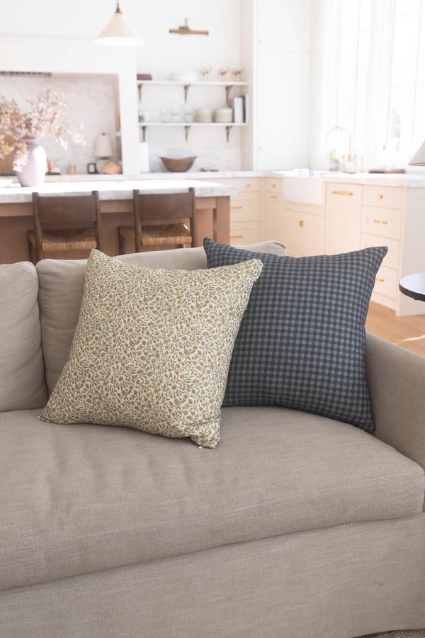 Two decorative pillows on a beige sofa: one with cream leaf pattern, one with charcoal gingham check.