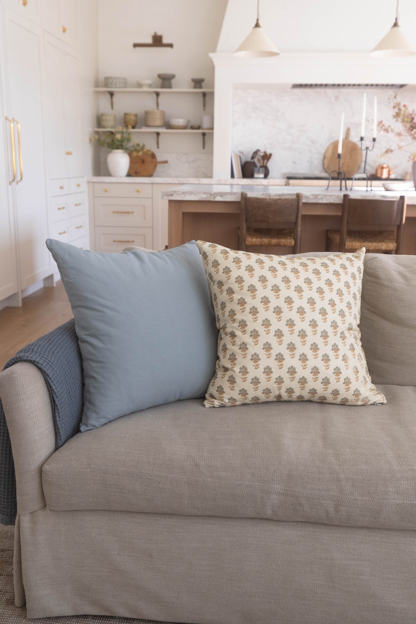Four decorative pillows in blue, patterned cream, and neutral tones arranged on a beige upholstered sofa.