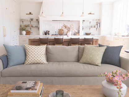 Dusty blue, cream, and gray decorative pillows arranged on a beige sofa in a bright kitchen and living room setting.