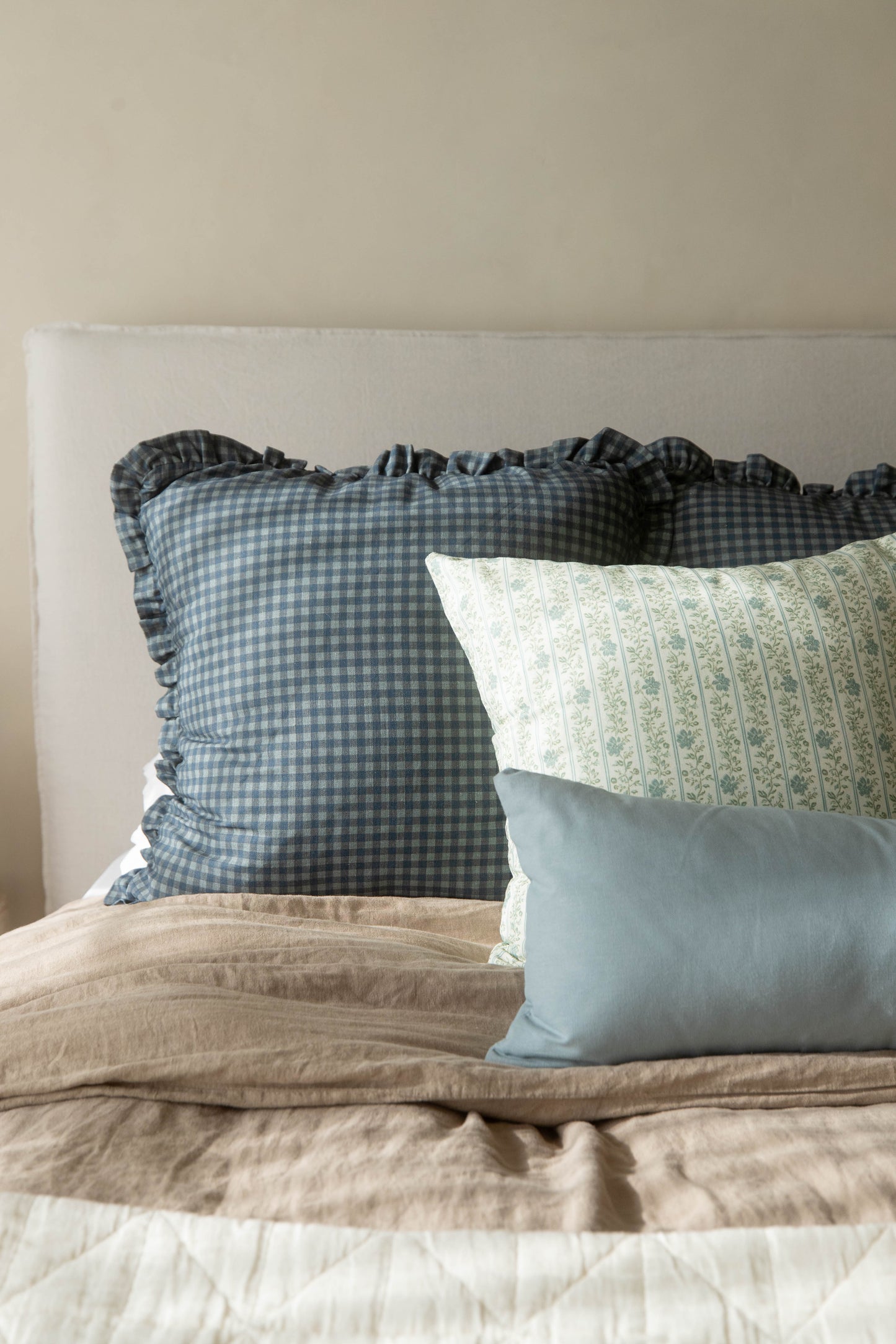 Dusty blue gingham pillow with ruffled edges displayed on a bed with patterned and solid pillows.