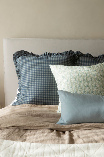 Dusty blue gingham pillow with ruffled edges displayed on a bed with patterned and solid pillows.
