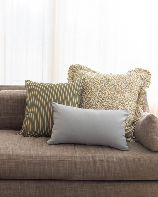 Light gray rectangular pillow with solid fabric displayed on beige sofa between striped and patterned accent pillows.