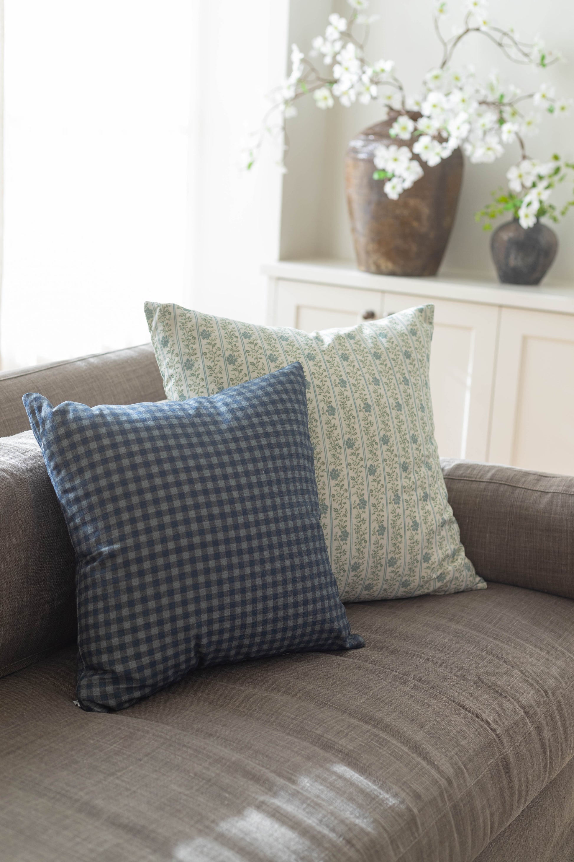 Two decorative pillows on a gray sofa: one with blue gingham pattern and one with green floral stripe design.